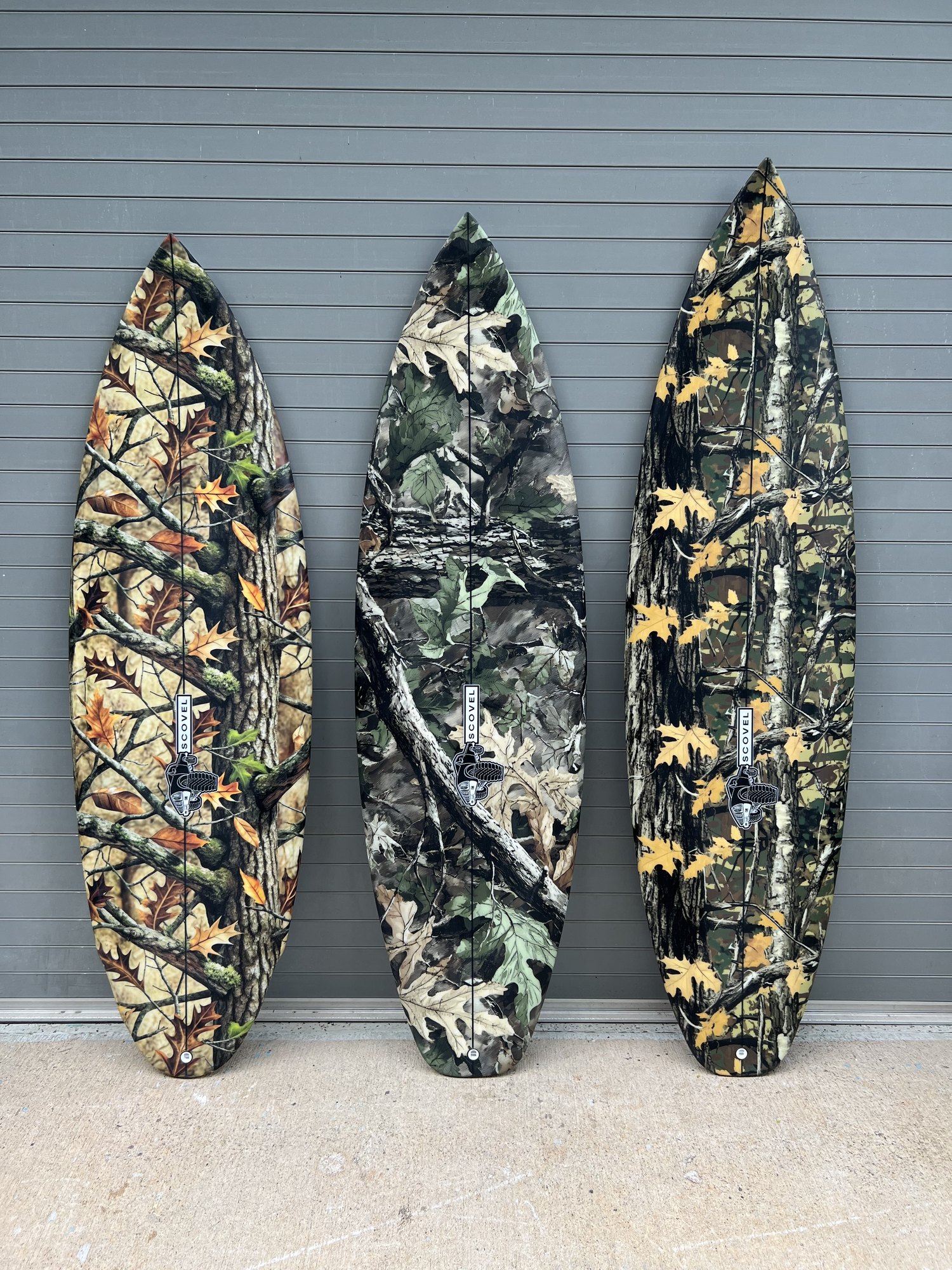 Three Scovel shortboards with forest camo leaf patterns laid against a grey wall